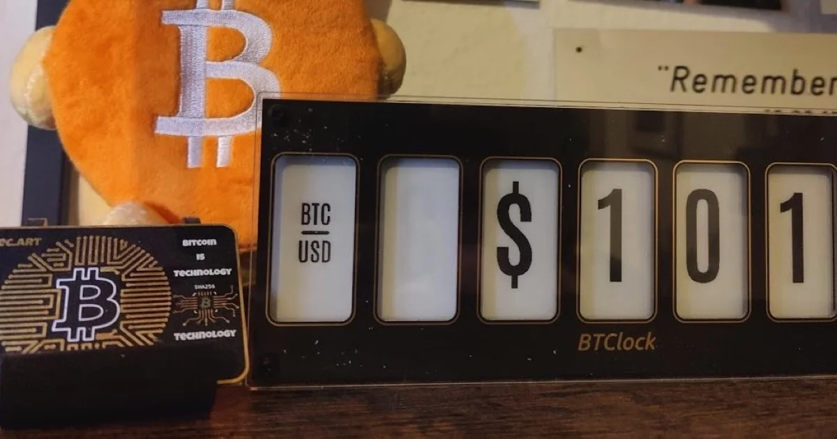 A BTClock desk display showing the BTC/USD price, next to a Bitcoin plush and a circuit card etched with the Bitcoin logo